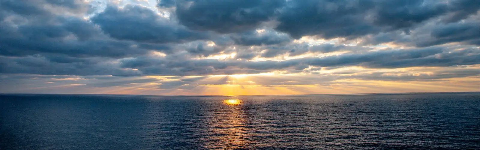Image with a sunrise above the ocean while reflecting in the water with rays coming through the clouds.