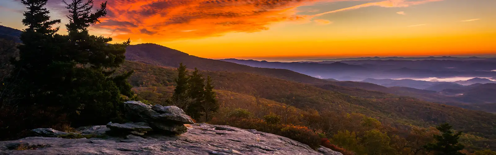 A panoramic view of a landscape with a rising sunrise with mountains and valleys in the foreground.