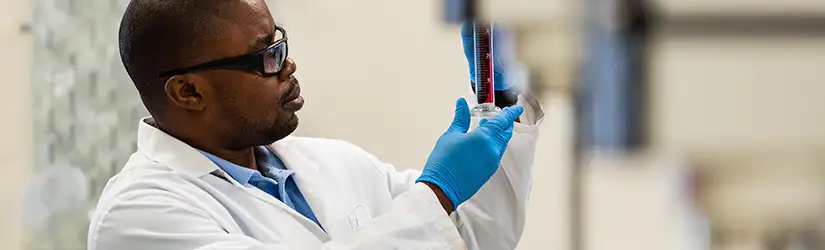 A person in a laboratory wearing a lab coat, safety glasses, and blue gloves inspecting a glass test tube, containing red fluid.