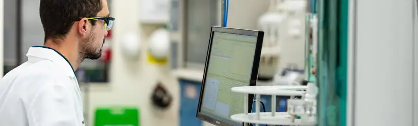 Man in a lab coat and safety glasses, viewing data on a monitor in a laboratory.