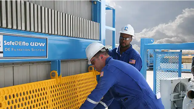 Two employees working with a TETRA SafeDEflo filtration system, while wearing safety equipment including white hard hats, protective eyewear, and blue overalls.