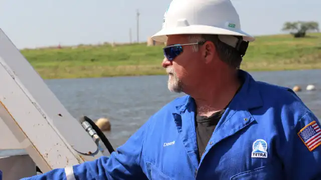 TETRA worker wearing a white safety helmet, protective eyewear, in a blue overall by a body of water.