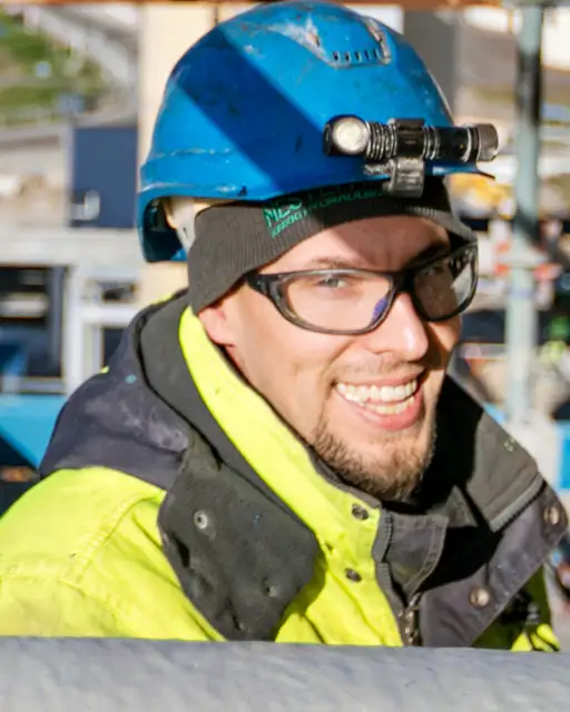 Työmaalla hymyilevä työntekijä, jolla on päässään sininen kypärä, suojalasit sekä kelta-musta takki.