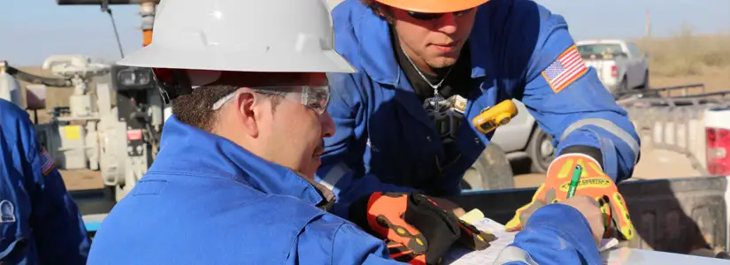 Two workers on a job site, in hard hats, safety glasses, and gloves going over plans on the back of a truck bed rail.