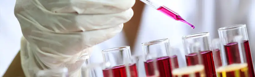 A hand wearing a white glove, holding a pipette to drop pink fluid into a test tube containing red fluid.