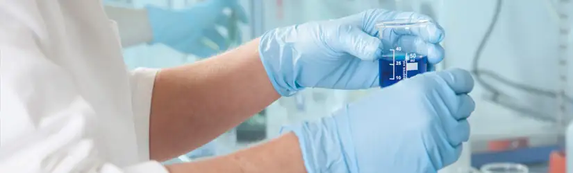 Person in a laboratory wearing a lab coat and blue gloves, holding a beaker containing blue liquid.