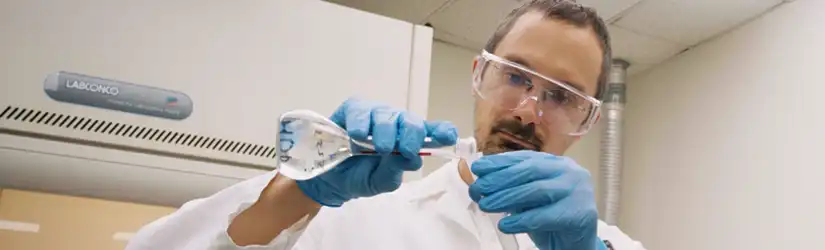 A TETRA employee working in a laboratory setting, holding a glass flask and conducting a fluids test while wearing safety goggles and blue nitrile gloves.