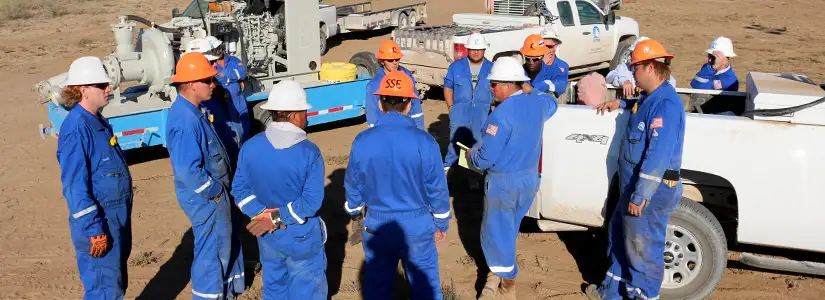 A group of workers gathering for a discussion by vehicles and equipment, wearing hard hats and overalls.