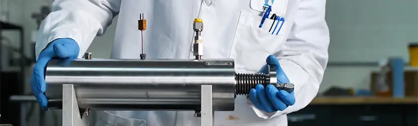 A person in a laboratory coat and blue gloves holding a metal cylinder tool for testing and product development.