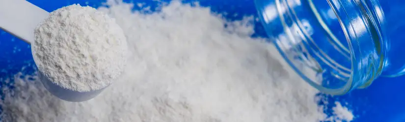 White dry salt powder material, with a scoop placed in a plastic measuring spoon, by a clear glass jar reseting on its side.